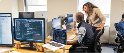 A woman leans over to assist a man working on code at a computer in a modern office. Multiple monitors display code, and the atmosphere is collaborative.