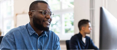 A man in glasses and a blue shirt smiles while working at a computer. Another person in the background works attentively in a bright office space.