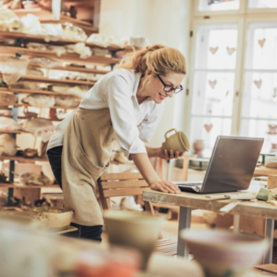 A woman in an apron working on a laptop in a pottery workshop.