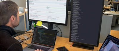 A bearded man sitting at a desk with a laptop and two monitors.