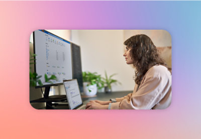 A woman working on a laptop with a larger monitor next to it in an office setting.