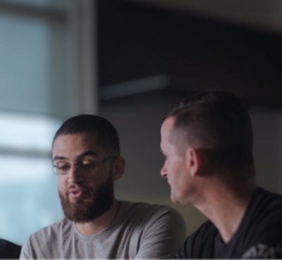 Two men, one with a beard, looking at a laptop screen together in a modern office setting with large windows.