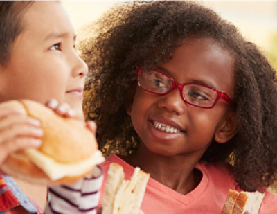 A young girl wearing glasses is looking at another girl holding a sandwich.
