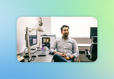 A man in a light blue shirt sitting at a desk with computer monitors, a lamp, and a coffee mug, in a brightly lit room.