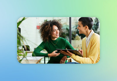 Two people are sitting at a table and smiling. One of them, wearing a green dress, holds a smartphone, while the other