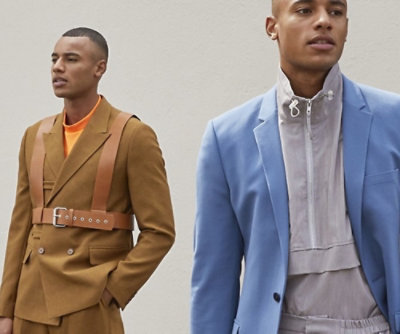 To menn i stilige antrekk mot en enkel bakgrunn. En har på seg en brun dress med en oransje skjorte og lærseler, den andre en blå dress med en høyhalset grå topp