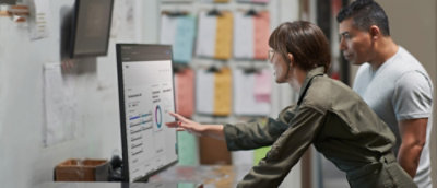 A woman pointing at a computer screen while a man in a grey shirt sits next to her at a desk.