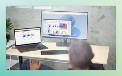 Person sitting at a desk with a large monitor displaying charts and graphs, and a laptop showing data