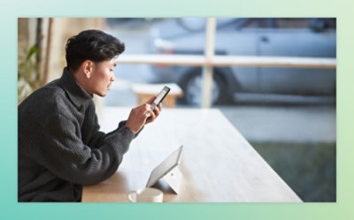 Person sitting at a table, holding a smartphone and looking at it, with a tablet on the table