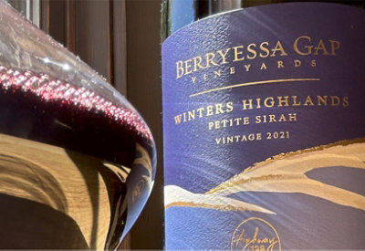 A wine glass filled with deep red wine sits beside a bottle labeled "Berryessa Gap Vineyards, Winters Highlands, Petite Sirah, Vintage 2021."