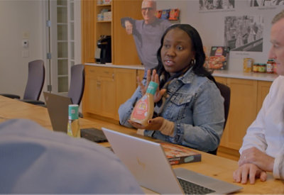A woman in a denim jacket presents a salad dressing bottle during a meeting, seated at a table with laptops. A collaborative, focused atmosphere.