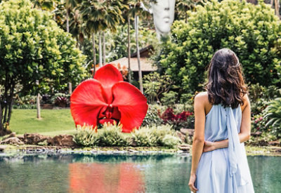 A woman in a flowing blue dress stands by a pond, facing a lush garden with a giant red flower sculpture and a serene outdoor ambiance.