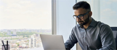 ノート PC を置いたデスクの前に座っているあご髭を生やしたメガネの男性。
