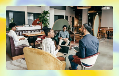 Four professionals in a modern office setting, engaged in a discussion with laptops. two are seated on sofas, two on armchairs.