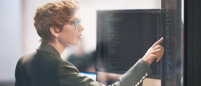 A person sitting at a desk and looking at a computer screen with a hand pointing at the screen.