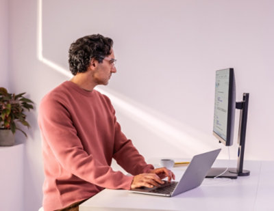 Man in a pink sweater works at a desk with a laptop and a monitor.