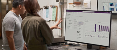 Two people reviewing a dashboard with charts on a computer monitor, with clipboards and another screen in the background.