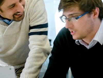 Two men sitting in front of laptops, smiling and looking at each other.