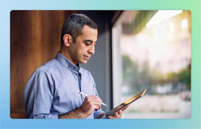 A man stands near a window, holding and writing on a tablet with a stylus, dressed in a blue button-up shirt.
