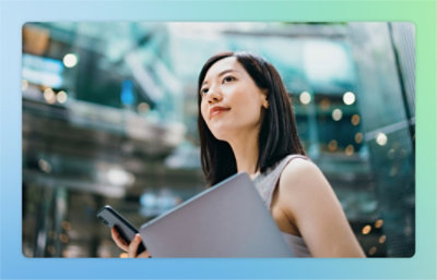 A woman holds a laptop and a smartphone, looking up, with a modern, glass-walled building in the background.