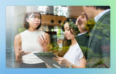 Three people in a business meeting. One woman speaks while the other woman focuses on a clipboard. 