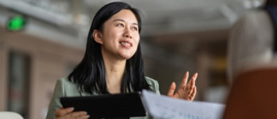 A woman with dark hair is holding a tablet and gesturing with her hand, while discussing something.