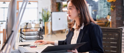 A woman in business attire working at a desk with a computer monitor, holding a document folder.