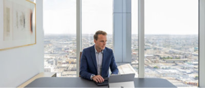 A man sitting at a desk by a large window, working on a laptop