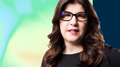 Person with long, wavy dark brown hair wearing a black top against a green‑blue gradient background