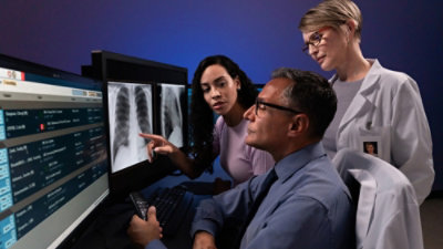 Three doctors examine a computer screen displaying an X-ray image, discussing findings collaboratively.