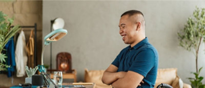 Smiling man in a blue shirt sits with arms crossed at a desk with a laptop, lamp, and plants. The room has a cozy, relaxed atmosphere.