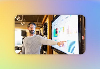 En mann i en grå genser peker på statistikk på en stor skjerm som viser fargerike grafer og diagrammer.