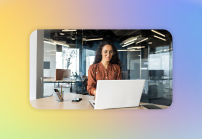 En kvinne med briller og langt krøllete hår jobber på en bærbar datamaskin i et moderne kontor.