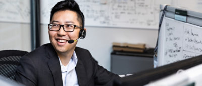 A man wearing glasses and a headset near a whiteboard with writing and text