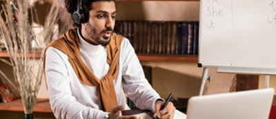 Man wearing headset teaching online, seated at a desk with laptop and whiteboard.