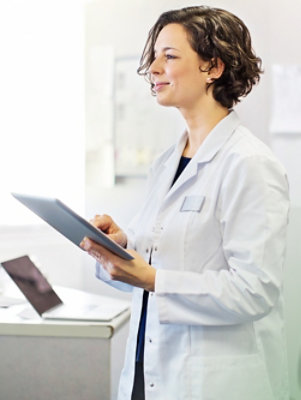 A woman in a lab coat smiles while holding a tablet in a bright office setting.