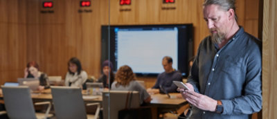 Person using a smartphone near a glass wall while a group works on laptops in a conference room with a large screen.