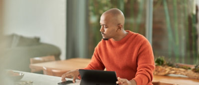 Person in an orange sweater working on a tablet at a wooden table in a bright room with large windows.