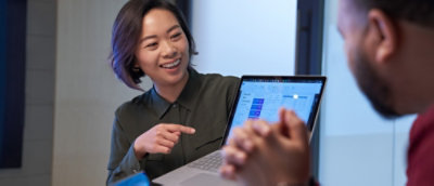 Person pointing at a laptop screen displaying a calendar and charts during a discussion with another individual.