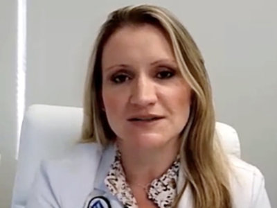 Woman with long blonde hair and a serious expression, wearing a white coat.