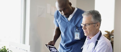 Two healthcare professionals review a medical scan on a tablet. One wears scrubs and the other has a stethoscope.