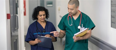 A man in green scrubs holding a folder and a woman in blue scrubs holding a clipboard.