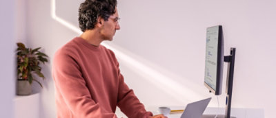 Man in a pink sweater works at a desk with a laptop and a monitor.