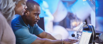 Two people look at a computer screen displaying a list or spreadsheet in a blue-lit office or lab environment.