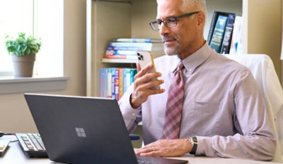 A man holding a phone and looking at a laptop with a potted plant nearby.