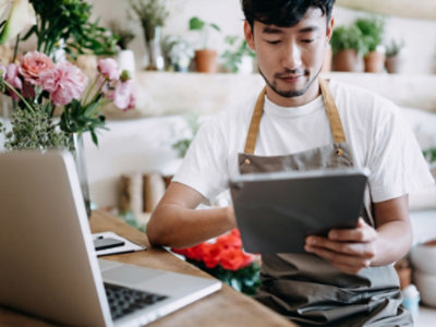 A man wearing an apron using a tablet while sitting in front of a laptop.