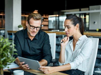 Two people looking at a tablet while sitting at a desk.