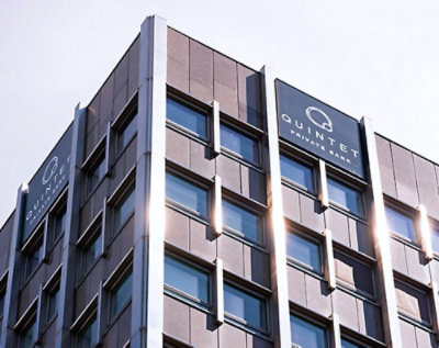 Grå moderne kontorbygning med "quintet"-logoet på toppen, under en klar himmel, hvor sollyset reflekteres i vinduerne.