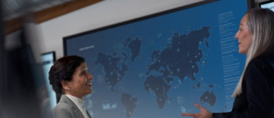 To kvinder, der har en samtale foran en stor computerskærm, der vises i baggrunden