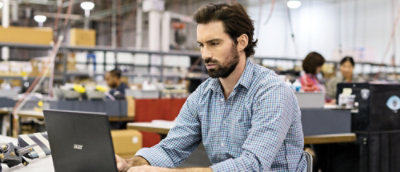 A bearded man sitting in a room looking at a laptop.